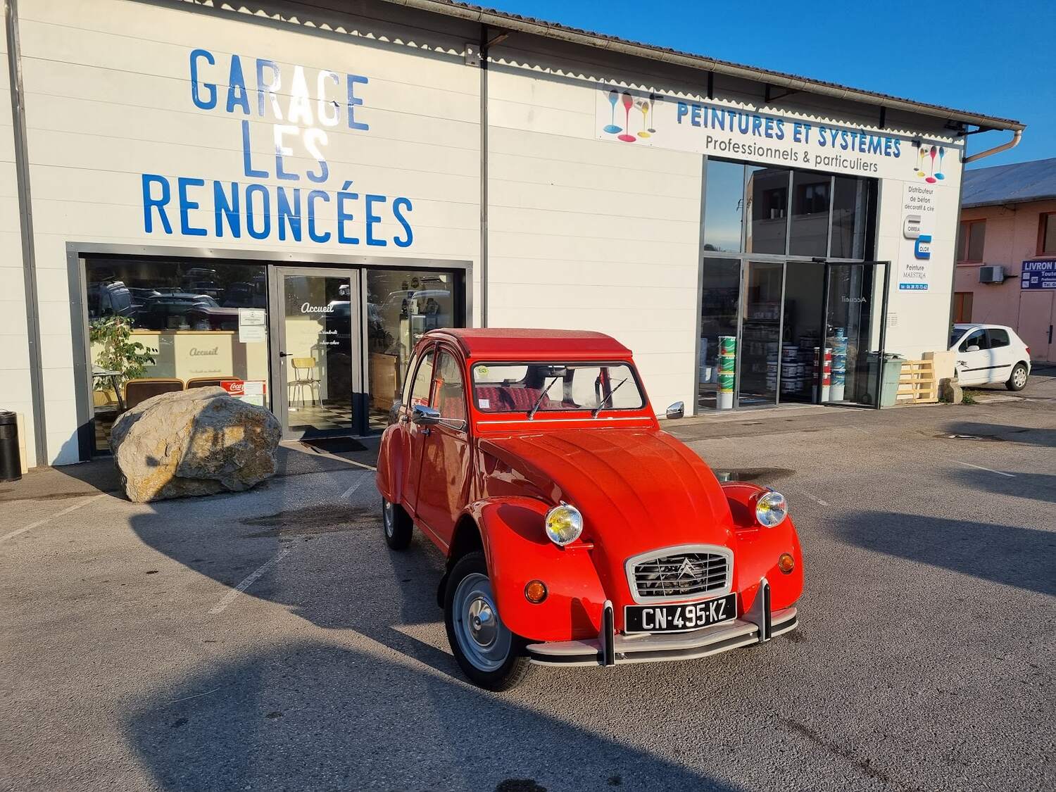 La façade blanche du Garage les Renoncées à Livron-sur-Drôme, son enseigne en lettres bleues, et une Citroën 2CV rouge garée devant